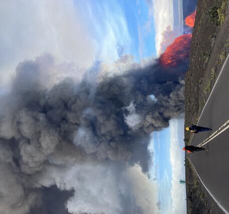 Color photograph of scientists walking on a closed road with volcanic eruption in the distance