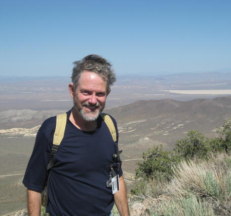 Man standing on a hill wearing a backpack with a view of sky and clouds in the background