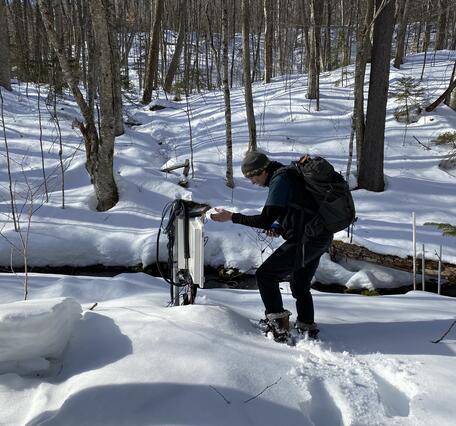 A man in the snowy woods looks at a data logger.