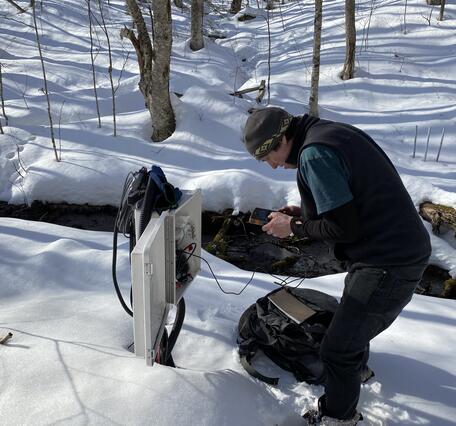 A man in the woods in the snow looks at a data logger.