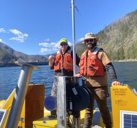 two males stand in safety gear and hats stand on a bright yellow floating platform in blue lake water on a sunny day