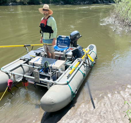 Man standing in an inflatable catamaran boat laden with equipment on the river bank 