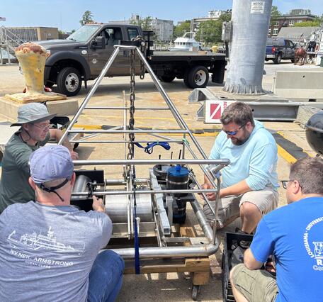 four people around a large piece of equipment outside by the water