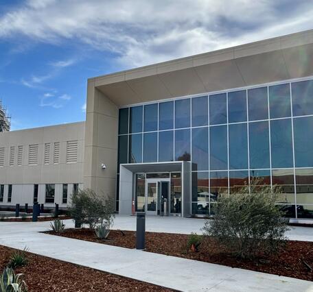 A rectangular beige building with a large, glass-fronted lobby is viewed at an angle. The building is surrounded by mulched landscaping and in the left background is a huge dirigible hangar under construction.