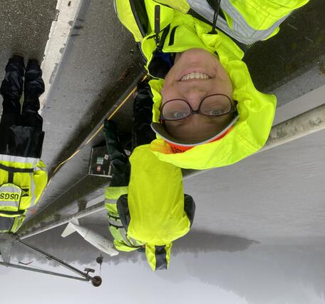 Bright yellow safety gear on USGS field crew stands out on a grey day as they work along a bridge over the river.