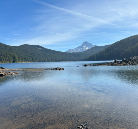 blue lake surrounded by rolling, forested foothills with mount Hood in the background. Blue sky day.