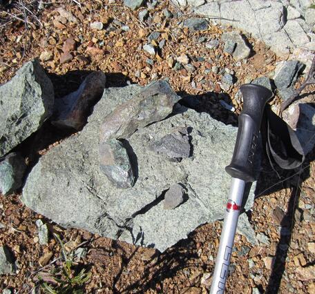 A greenish boulder with several shiny green rock fragments rests on pebble-strewed ground. The boulder is mottled with several colors of green minerals. The grip end of a hiking pole rests on the boulder to give a sense of scale. 