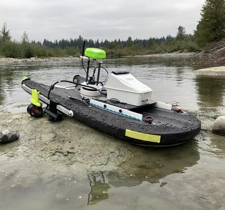 A small USV sits on a shallow riverbank, equipped with sensors, a propulsion unit, and a compact central hull