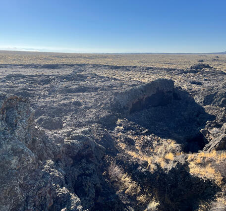 fissure in dark rock with dry grass, and dry grassy landscape in the distance under blue sky