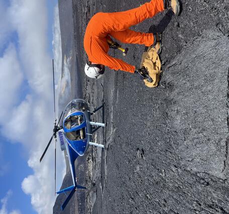 Color photograph of geologist collecting sample of lava flow with helicopter in background