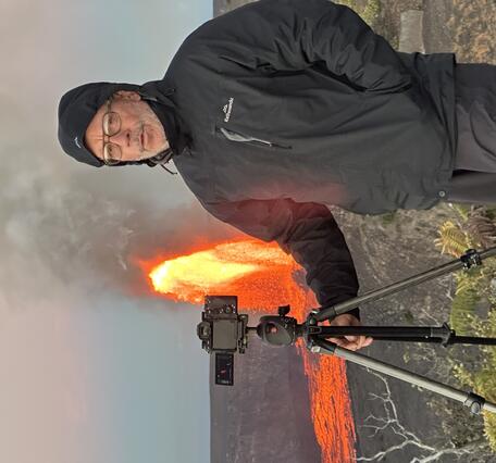 Color photograph of scientist bundled up with camera on tripod next to him and lava fountain erupting in the background