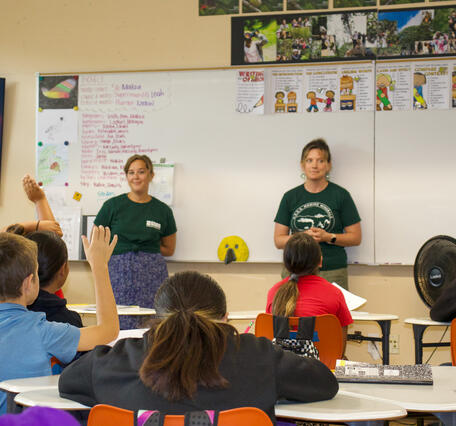 Two people stand at the front of a classroom of children. 