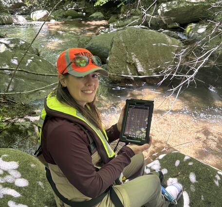 A biologist wearing an orange hat and yellow PFD, sits by a stream holding a tablet.