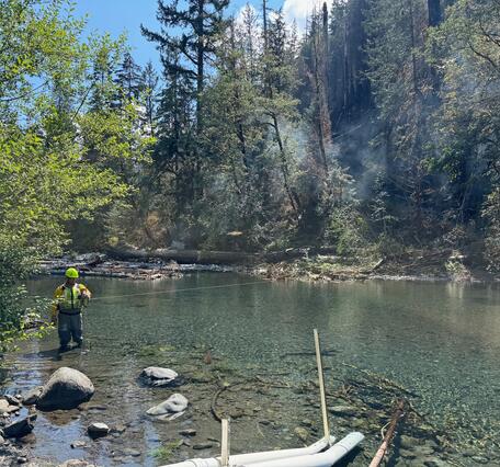 Technician stands in river measuring flow near fire