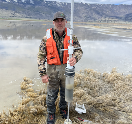 man on shallow lake shore holding a long, clear tube with a handle or frame at the top and a valve or stopper at the bottom