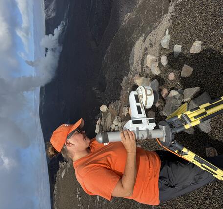 Color photograph of field engineer servicing a livestream camera with volcanic vents degassing in the background