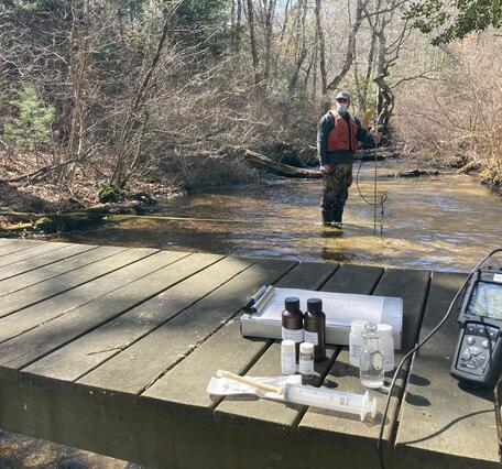 A man stands in a shallow river with a flow meter in the background and water quality equipment in the foreground on a dock.