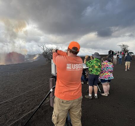 Color photograph of scientist setting up equipment with visitors viewing a volcanic eruption nearby