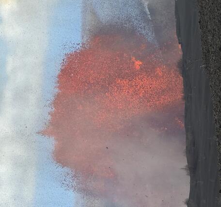 Color photograph of lava fountain during the day time with crater rim in the foreground