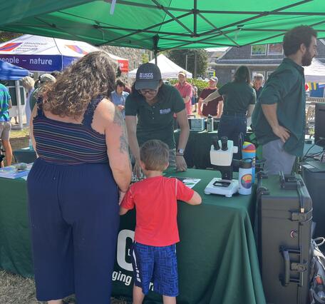 person behind USGS booth talking to adult and child outside under tent