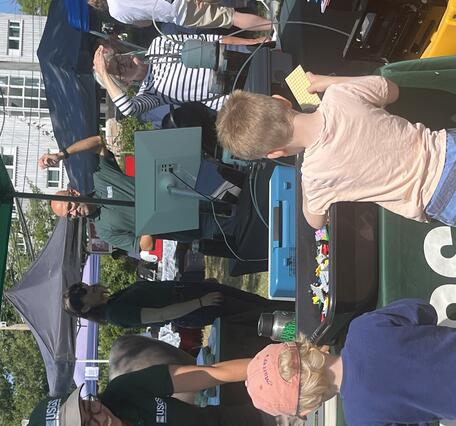Person in USGS hat and shirt at USGS booth outside smiling at kids playing with our Legos