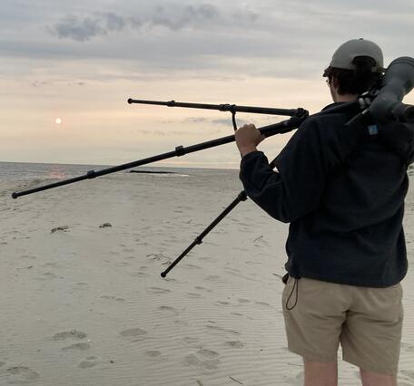 A man stands on a beach facing away from the camera holding a spotting scope