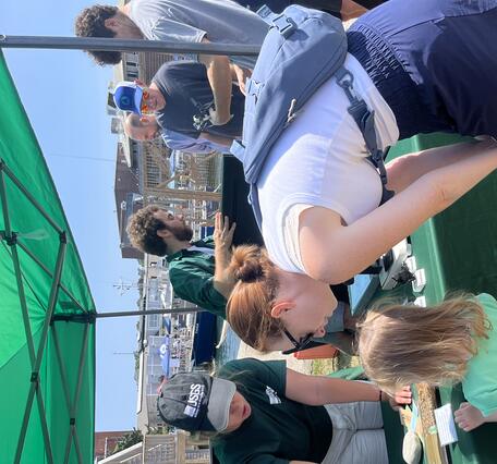 woman showing adult and child a replica sediment core on table outside under green tent