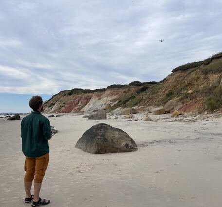 person standing on sandy beach with control in hand looking at drone in the sky over cliffs