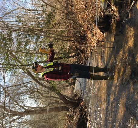 A man stands in a shallow stream with a flow meter.