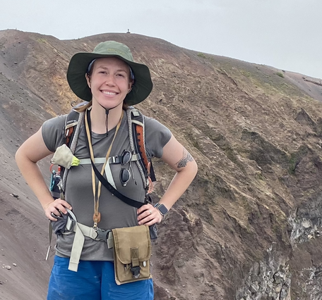 A young female geologist stands on a volcanic crater rim with her hands on her hips, wearing a gray shirt and green hat. 