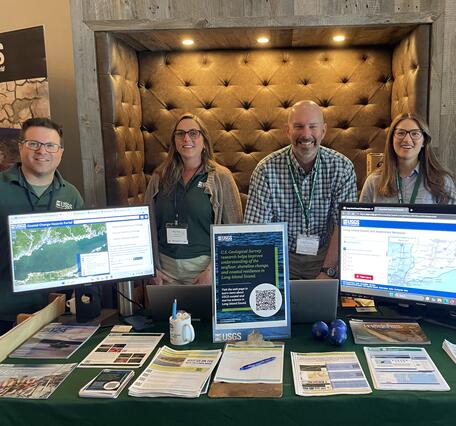 Group of USGS scientists smiling for photo behind booth with handouts and two computer monitors displaying maps