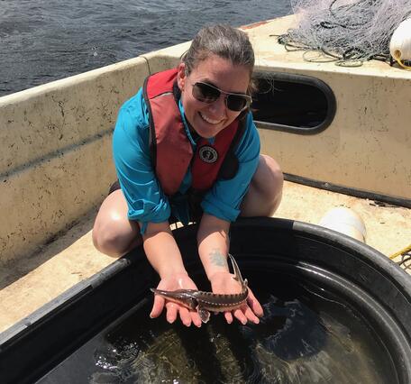 A person in a blue shirt and orange life jacket is on a boat, leaning over a tub of water, holding a small fish in their hands