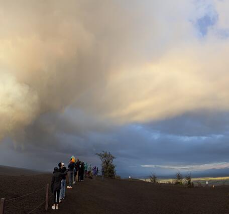 Color photograph of lava fountain in crater, visitors and rainbow on the rim