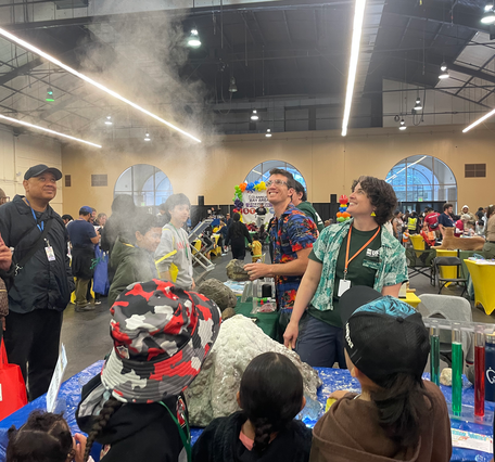 This photograph shows a model volcano sitting on a table surrounded by people. The model volcano has a plume of fine material suspended in the air above it, indicating it erupted immediately before the photo was taken. In front of the table, children participants face away from the camera looking at the volcano and the plume. Behind the table, animated scientists in brightly colored shirts are smiling and looking up at the plume.
