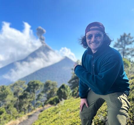 male scientist wearing a backwards hat and sunglasses, pointing towards and erupting volcano in the background.