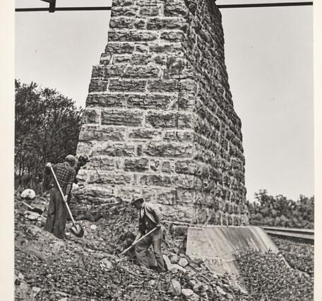 Black and white photo of men digging at the foot of an old bridge