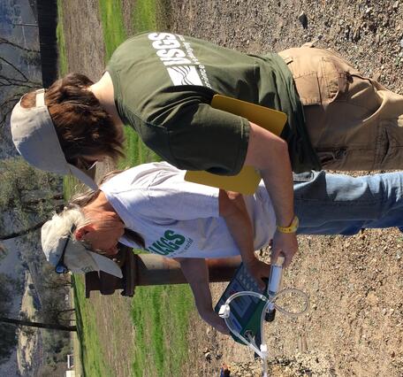 Two USGS scientists  wearing USGS shirts and baseball caps stand next to a rusting wellhead pipe and concrete well casing on a gravel field. One holds a gas analyzer with tubing; the other takes notes on a yellow clipboard. A green valley and low mountain ridges rise in the background under a clear blue sky.