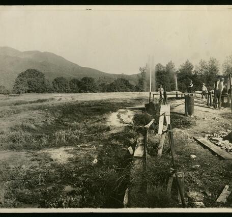 A sepia-toned historical photograph shows an open field with people standing near a row of wooden-framed well structures and pipes. A thin jet of gas or water sprays upward from two of the wells. Forested hills and a mountain rise in the background. 