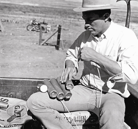 Man in white button-down shirt sitting on bags of explosives, assembling dynamite sticks used as detonator