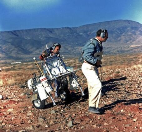 Two men wearing jackets and headsets pulling a piece of equpment up a moderate slope in the desert southwest