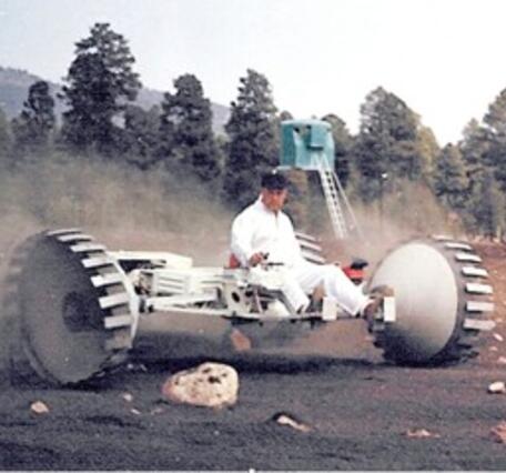 Early lunar roving vehicle prototype with large wheels and an open seat, being driven by a man in a white jumpsuit