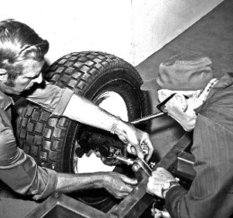 A black and white photo of two men working on the suspension of an open chassis vehicle