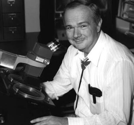 Gene Shoemaker wearing a white button-up shirt and a bolo tie, sitting at a microscope and looking at the camera