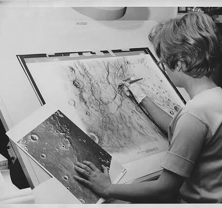 A woman with short hair sits at an inclined desk, adding detail to a map of the lunar surface with an airbrush