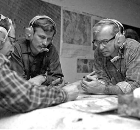 Three men with headsets are hovering over maps and appearing to discuss observations