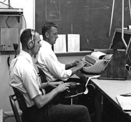 Two men in white shirts sitting at typing machines: a typewriter and a stenograph