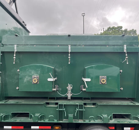 Rear view of a green industrial trailer or tank body, mounted on a flatbed truck chassis. The tank has two arched access doors with round latch handles and gauge/valve fittings, flanked by turnbuckle tensioners along the top edge. Cables run between the two door latches. Below, the flatbed frame shows tie-down hooks and chain slots. 