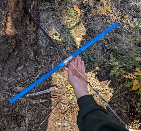a hand holds an X-shaped tool, with one end of the tool stuck into a tree, leaves and grasses on the ground