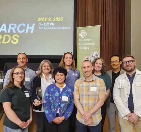 10 people stand and smile in front of a light wood stage with a projector screen saying: 2026 Research Awards
