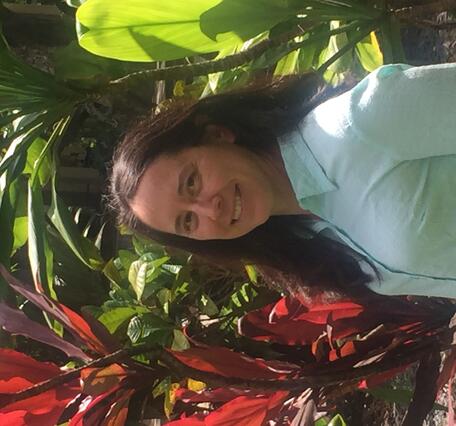 A profile picture of Janet Cushing in Hawaii. A woman with long dark hair is standing in front of bright, green and red tropical foliage while wearing a blue button up shirt. She is smiling at the camera. 